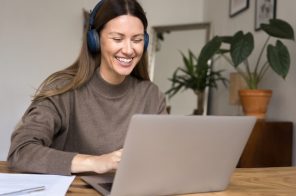 Mulher com blusa marrom e headphones azuissentada à mesa do computadorsorrindo enquanto pesquisa como trocar de internet no notebook.