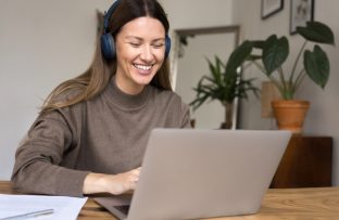 Mulher com blusa marrom e headphones azuis, sentada à mesa do computador, sorrindo enquanto pesquisa como trocar de internet no notebook.