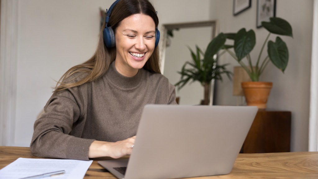 Mulher com blusa marrom e headphones azuis, sentada à mesa do computador, sorrindo enquanto pesquisa como trocar de internet no notebook.