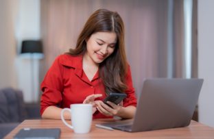 Mulher com camisa vermelha sentada à escrivaninha, com a sala de estar ao fundo, sorrindo de cabeça baixa enquanto navega no celular conectado ao Plano Controle da Desktop. Sobre a mesa, há um notebook cinza, uma caneta e uma caneca branca.