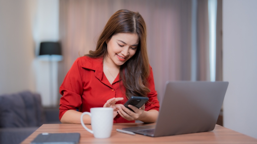 Mulher com camisa vermelha sentada à escrivaninha, com a sala de estar ao fundo, sorrindo de cabeça baixa enquanto navega no celular conectado ao Plano Controle da Desktop. Sobre a mesa, há um notebook cinza, uma caneta e uma caneca branca.