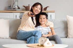 Mãe e filho sentados no sofá, abraçados e sorrindo, enquanto a mãe aponta o controle para a TV que exibe streaming infantil durante as férias.