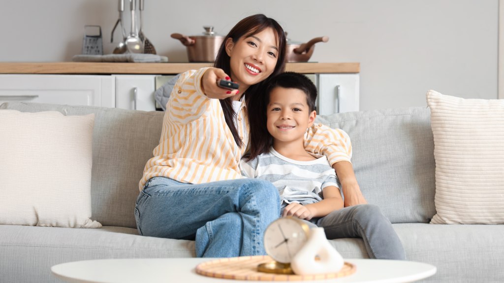 Mãe e filho sentados no sofá, abraçados e sorrindo, enquanto a mãe aponta o controle para a TV que exibe streaming infantil durante as férias.