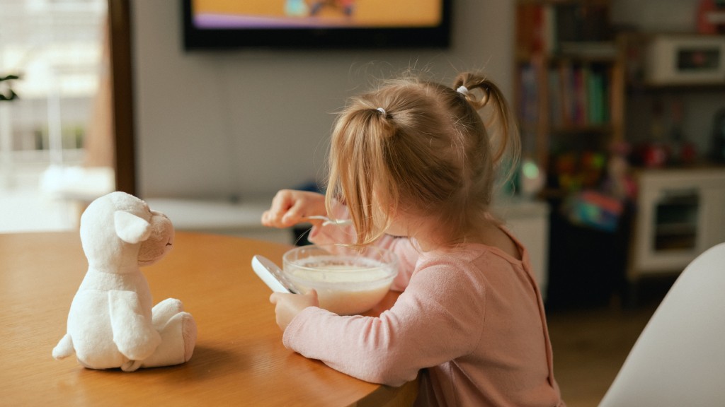 Menina com blusa rosa e chiquinhas no cabelo sentada à mesa de jantar, comendo leite com cereais ao lado de um cachorrinho de pelúcia, enquanto uma TV à direita exibe um desenho em streaming, ilustrando dicas sobre o que fazer nas férias.