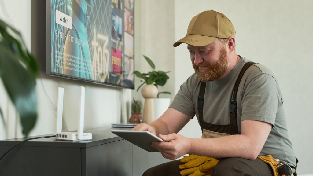 Técnico de internet com camiseta e boné verdes e avental preto, agachado próximo a um roteador branco na sala de estar, com um tablet na mão enquanto instala internet fibra óptica.