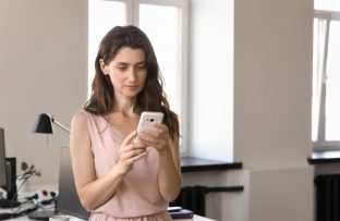 Mulher com vestido rosa claro de pé no escritório segurando celular enquanto aprende como gravar a tela do celular.