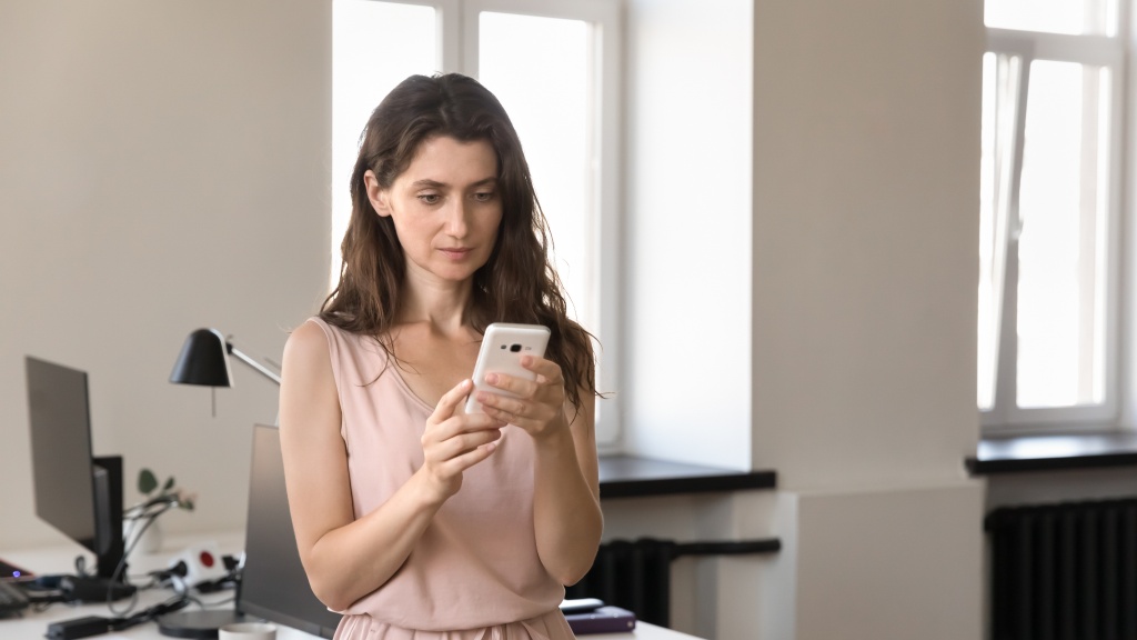 Mulher com vestido rosa claro de pé no escritório segurando celular enquanto aprende como gravar a tela do celular.