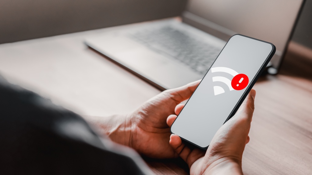 Mãos segurando um celular que exibe o ícone de Wi-Fi com uma exclamação em destaque vermelho, representando queda de sinal Wi-Fi. Ao fundo, desfocado, há um notebook sobre a mesa.