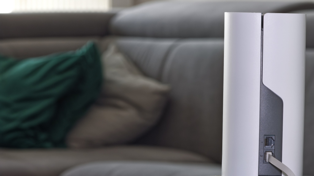 Roteador mesh branco sobre mesa lateral de uma sala de estar, com sofá cinza desfocado ao fundo.