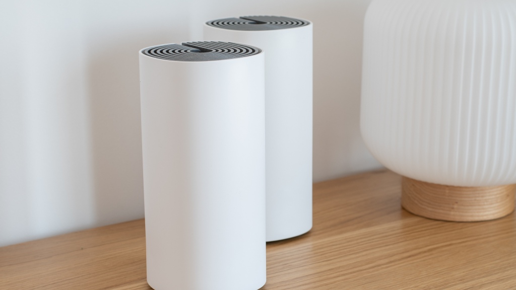 Dois roteadores mesh brancos sobre uma mesa de madeira, ao lado de um objeto decorativo dourado e branco.