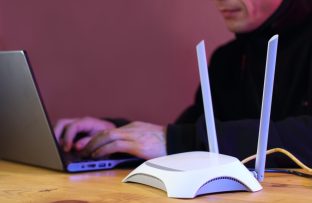 Destaque para um roteador mesh com duas antenas sobre uma mesa de madeira, com um homem de blusa preta desfocado ao fundo navegando no notebook e parede iluminada em tom rosa.