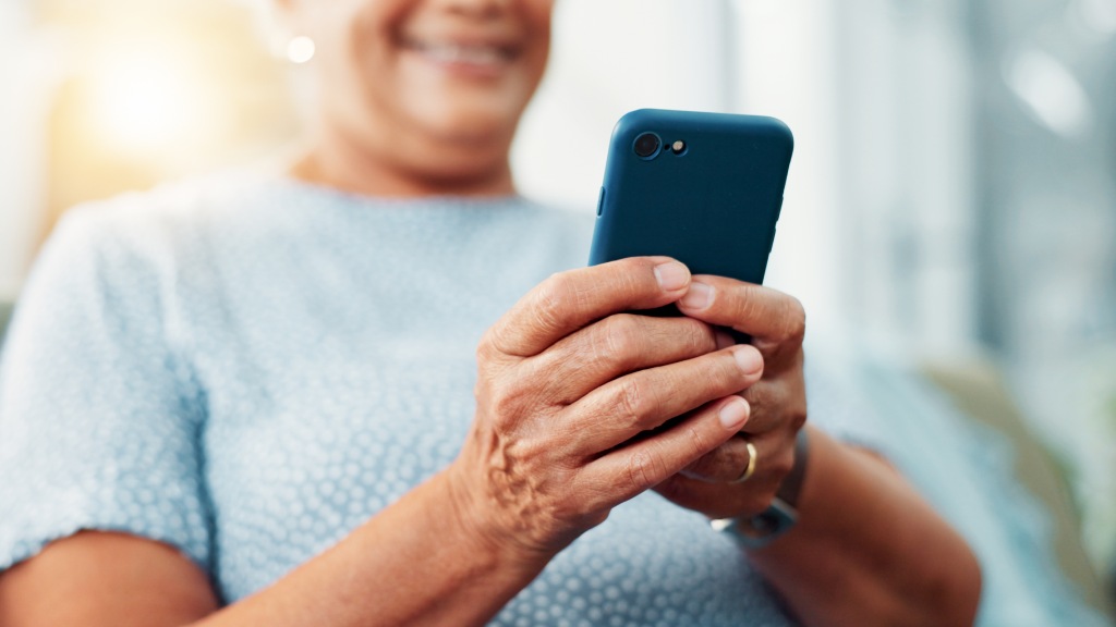 Mulher idosa com blusa azul de bolinhas brancas, sorrindo, segurando um celular em destaque, representando um aparelho funcional graças à rotina de limpeza da memória.