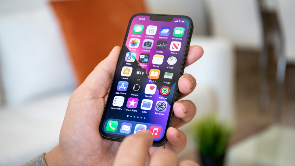 Mãos segurando um iPhone que exibe a tela inicial, com fundo em tons de roxo e vários aplicativos, representando o uso da nuvem para liberar espaço no celular.
