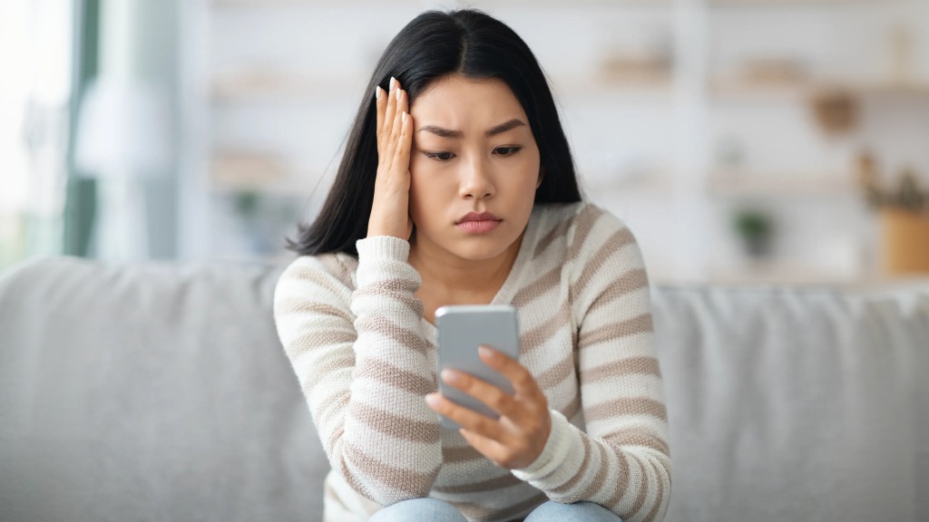 Mulher jovem com blusa listrada branca e marrom sentada no sofá, com uma das mãos no rosto, séria e preocupada, tentando resolver um celular perdido pelo aplicativo Celular Seguro.