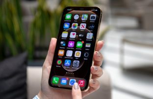 Mãos segurando celular iPhone, que exibe a Tela de Início do iPhone, representando a dúvida de como desativar aplicativos em segundo plano no aparelho.