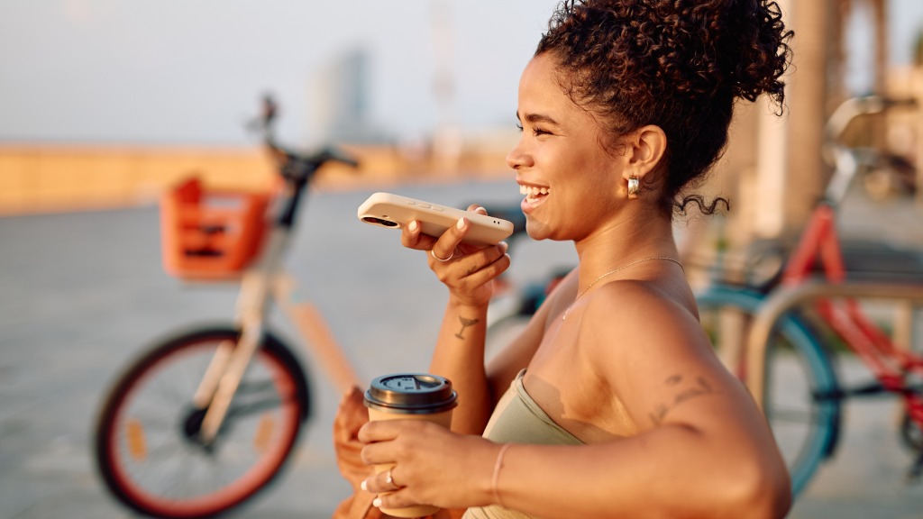 Mulher jovem ao ar livre, sorrindo enquanto grava um áudio pelo celular e segura um copo de café com a outra mão, representando uma pessoa que economiza dados móveis. Ao fundo, duas bicicletas desfocadas.