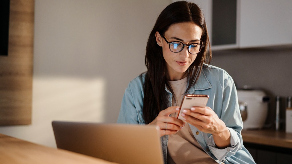 Mulher com camisa jeans de botões e camiseta bege usando óculos de grau redondos, sentada à mesa de notebook com celular na mão enquanto desativa aplicativos em segundo plano no aparelho.