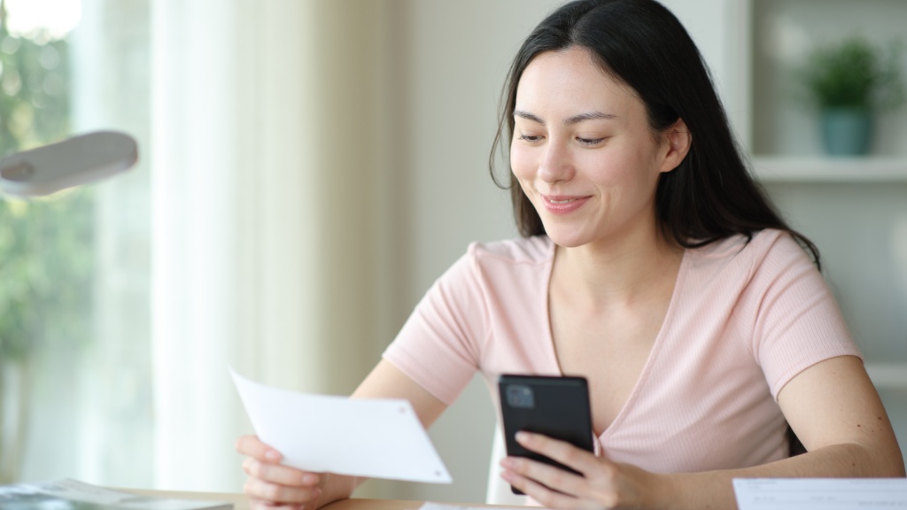 Mulher jovem com blusa rosa clara sorrindo sentada à mesa de trabalho com celular numa mão e boleto na outra usando Pix automático para pagar fatura.