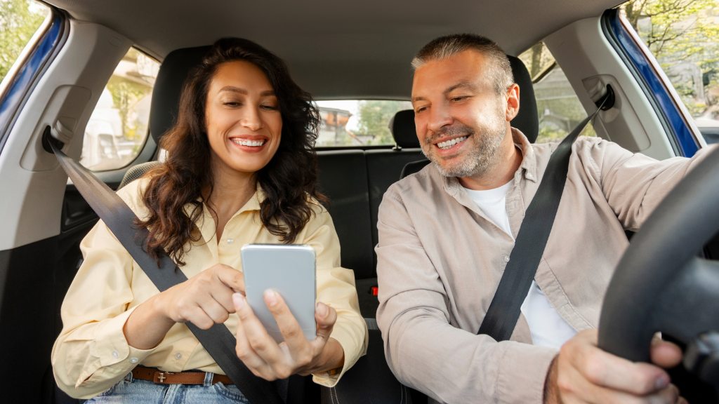 Casal no carro, ambos sorrindo: ela segura o celular e aponta para a tela, enquanto ele, com as mãos no volante, olha para o aparelho, representando usuários que sabem economizar dados móveis.