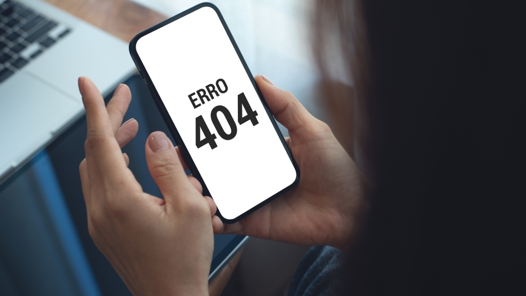 Mãos segurando celular, que exibe na tela o status "Erro 404" após acesso a uma página indisponível.