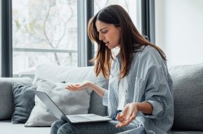 Mulher jovem com camisa de listras brancas e cinzas sentada no sofá, com notebook no colo e expressão de insatisfação por problemas de internet.