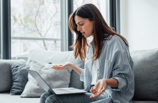 Mulher jovem com camisa de listras brancas e cinzas sentada no sofá, com notebook no colo e expressão de insatisfação por problemas de internet.