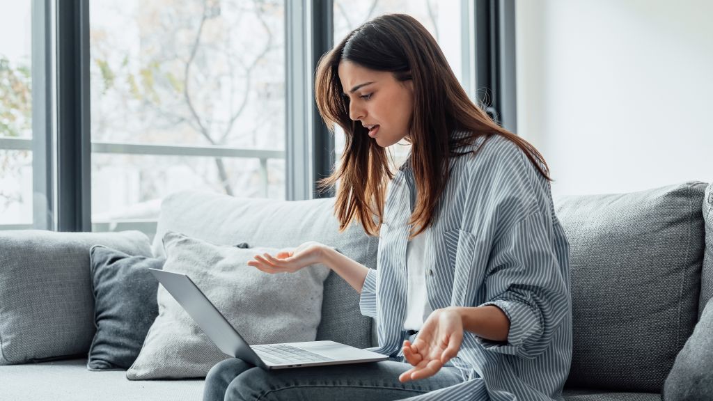 Mulher jovem com camisa de listras brancas e cinzas sentada no sofá, com notebook no colo e expressão de insatisfação por problemas de internet.