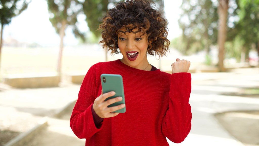 Mulher jovem com blusa vermelha ao ar livre, segurando o celular com o braço levantado e punho cerrado, empolgada enquanto navega com sinal de alta qualidade da Desktop Internet.