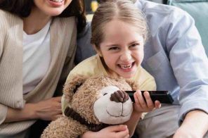 Família sorrindo no sofá enquanto a criança segura um controle remoto e um urso de pelúcia, assistindo a programação da HBO Max.