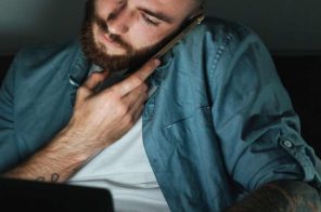 Homem jovem sentado na cama, em um quarto escuro, com notebook no colo enquanto fala ao celular, representando a dúvida sobre como colocar o Google no modo escuro.