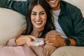 Casal no sofá, com o rapaz de blusa verde sentado e a moça de blusa marrom deitada sobre ele, ambos sorrindo enquanto ela segura o controle apontado para a TV que exibe conteúdo em um aplicativo de streaming.