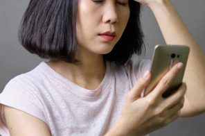 Mulher oriental com blusa rosa claro sentada, segurando o celular, com a outra mão na testa, olhos fechados e expressão de frustração devido à bateria viciada do aparelho.