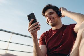 Homem jovem com camisa vermelha, usando headphones e segurando o celular ao ar livre, aproveitando um ótimo sinal de internet móvel no celular.