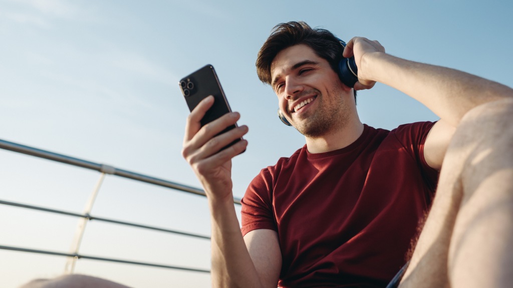 Homem jovem com camisa vermelha, usando headphones e segurando o celular ao ar livre, aproveitando um ótimo sinal de internet móvel no celular.
