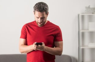 Homem de camiseta vermelha em pé, com a cabeça baixa, olhando para o celular enquanto tenta entender como economizar dados móveis.
