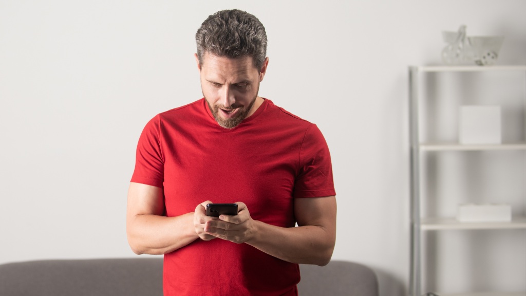 Homem de camiseta vermelha em pé, com a cabeça baixa, olhando para o celular enquanto tenta entender como economizar dados móveis.