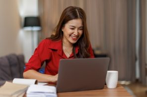 Mulher sorridente com camisa vermelha sentada à mesa de trabalho na sala de estar, usando o notebook com agilidade, representando bom desempenho da memória RAM.