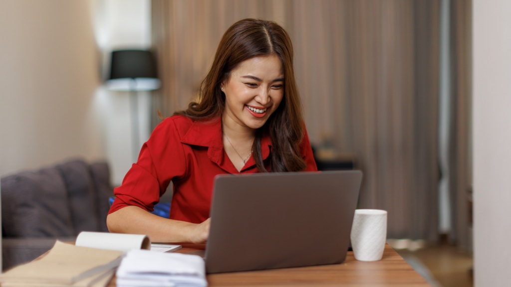 Mulher sorridente com camisa vermelha sentada à mesa de trabalho na sala de estar, usando o notebook com agilidade, representando bom desempenho da memória RAM.