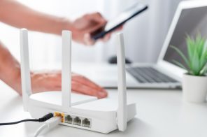 Modem de internet branco em bancada ao lado de notebook e vasinho de planta, com pessoa apoiada com uma das mãos enquanto conecta o celular à internet.