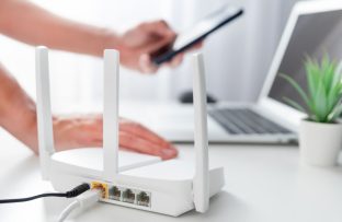 Modem de internet branco em bancada ao lado de notebook e vasinho de planta, com pessoa apoiada com uma das mãos enquanto conecta o celular à internet.
