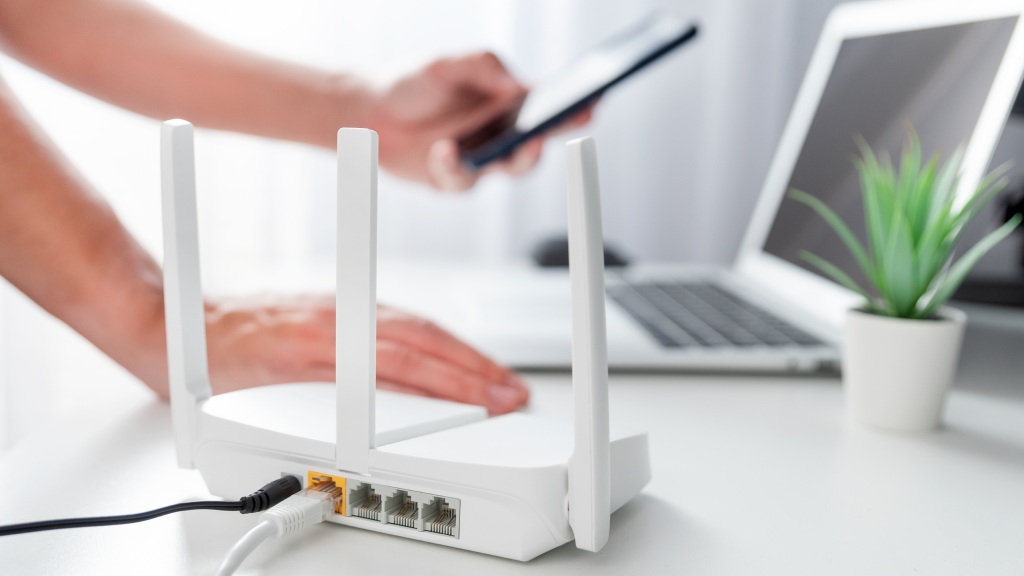 Modem de internet branco em bancada ao lado de notebook e vasinho de planta, com pessoa apoiada com uma das mãos enquanto conecta o celular à internet.
