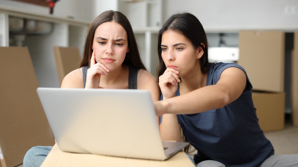 Duas moças sentadas, uma com blusa azul e outra com blusa cinza, com expressão de dúvida enquanto acessam o histórico do navegador no notebook.