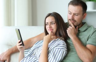 Casal sentado no sofá: ele com camisa polo verde e ela com blusa branca com listras azuis, ambos com o dedo sobre a boca; ela segura o celular enquanto ambos demonstram dúvida sobre o que fica salvo no histórico do navegador.
