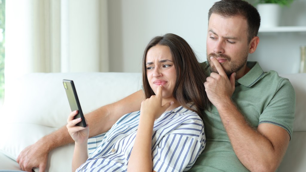 Casal sentado no sofá: ele com camisa polo verde e ela com blusa branca com listras azuis, ambos com o dedo sobre a boca; ela segura o celular enquanto ambos demonstram dúvida sobre o que fica salvo no histórico do navegador.