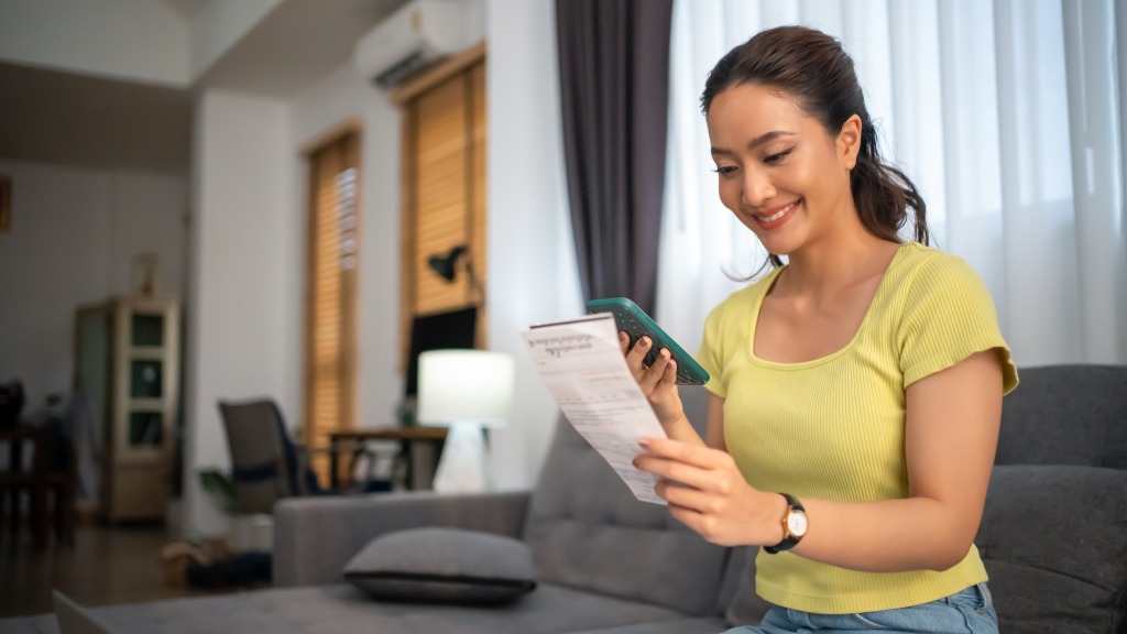 Mulher com blusa amarela, sorrindo, sentada no sofá enquanto escaneia um código Pix de boleto com a câmera do celular, habilitando o Pix automático pelo app.
