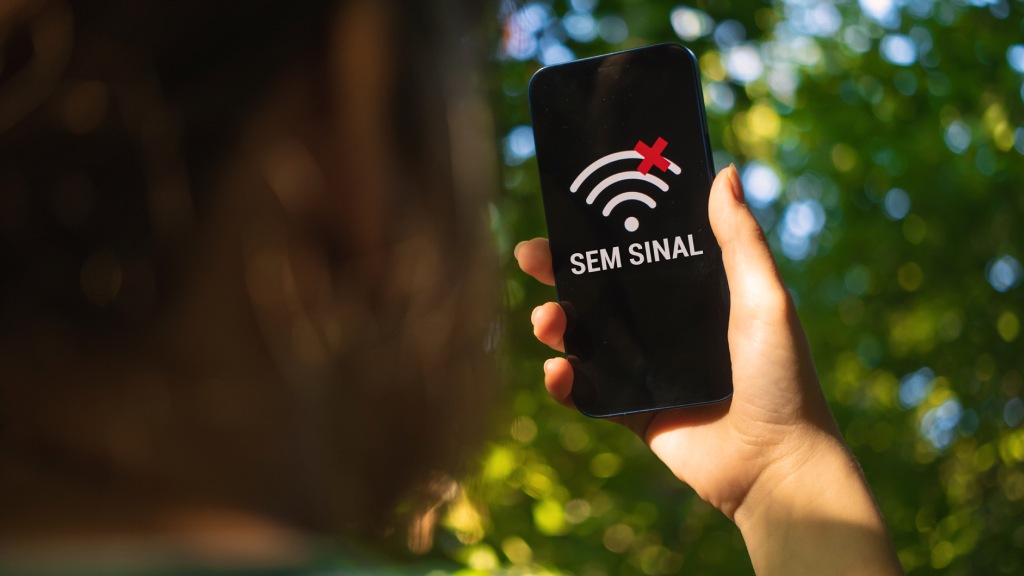 Mão segurando celular ao ar livre, com a tela exibindo o ícone de sinal Wi-Fi com um “X” vermelho sobreposto e, abaixo, o texto “Sem sinal”, representando problemas no sinal do celular.