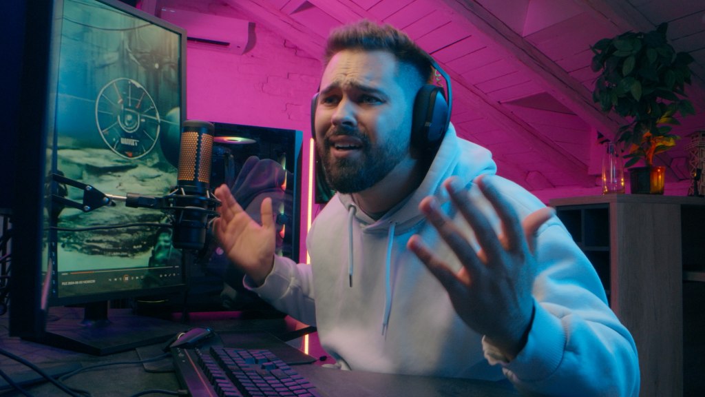 Rapaz gamer com moletom cinza usando headphones, com as mãos levantadas e expressão de decepção por lag causado por problemas de internet durante o jogo online.