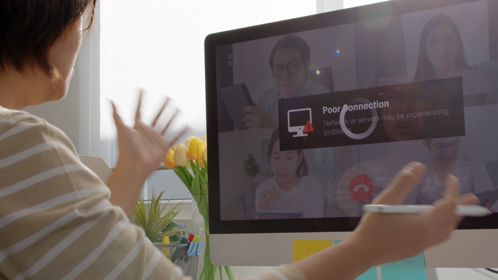 Mulher com as mãos levantadas em sinal de indignação sentada em frente ao monitor do PC que exibe o ícone de carregamento por problemas de internet durante uma videochamada.