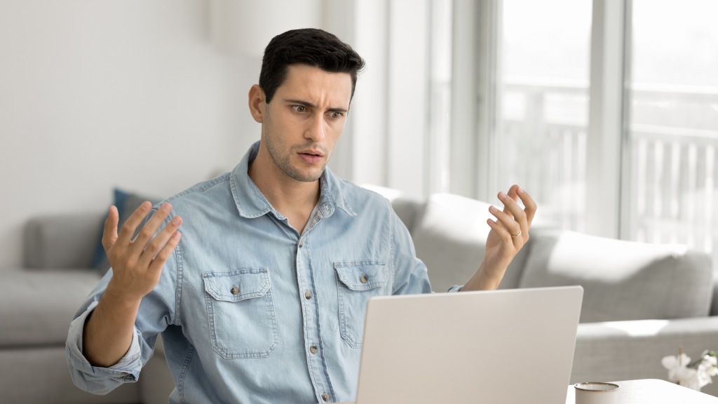 Homem jovem com camisa jeans, sentado à mesa da sala em frente ao notebook, com as mãos levantadas e expressão de frustração por problemas de internet no dispositivo.