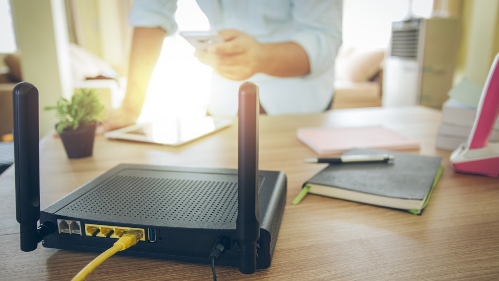 Modem preto com cabo amarelo conectado na porta traseira sobre bancada com cadernos de anotações, caneta, vaso de planta e tablet, com pessoa ao fundo com celular na mão.